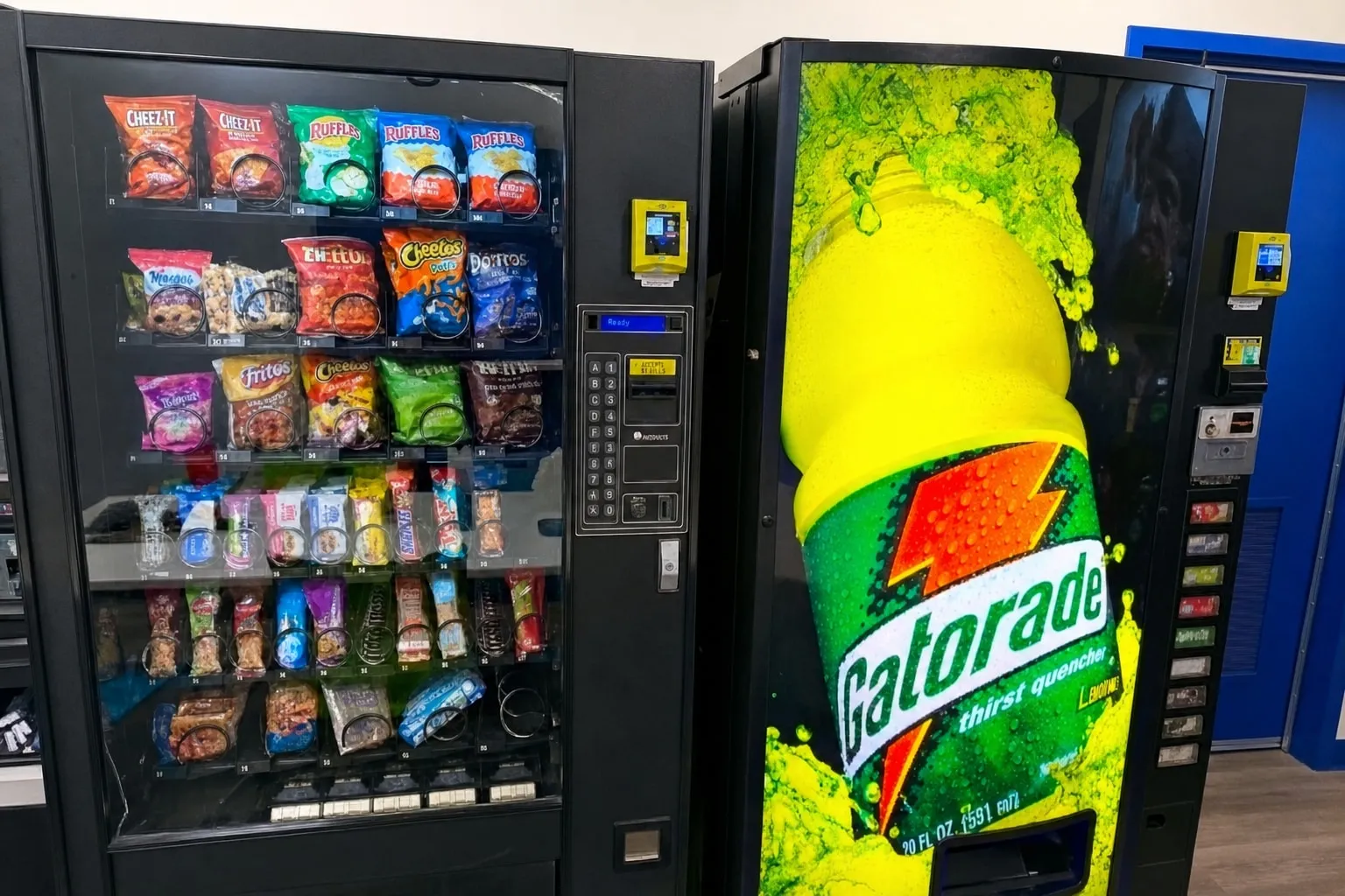 Snack vending machine and a sports-drink machine installed on a warehouse floor next to the safety board, serving every shift around the clock